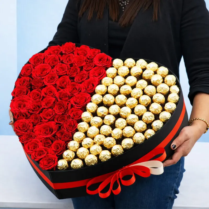 heart-shaped-box-of-red-roses-with-ferrero-2026-01-13 A woman holds a heart-shaped box filled with Ferrero chocolates and by 50 red roses, symbolizing love and affection.