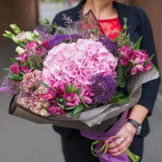 A women holding a bouquet of large pink hydrangea, complemented by various shades of purple, pink, and white flowers.