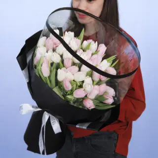 A person holding a large bouquet of pink and white tulips wrapped in black paper with a clear plastic cover