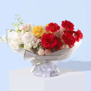A bouquet featuring mixed roses,carnations, and green foliage, wrapped in white paper with a gold trim and a white ribbon.