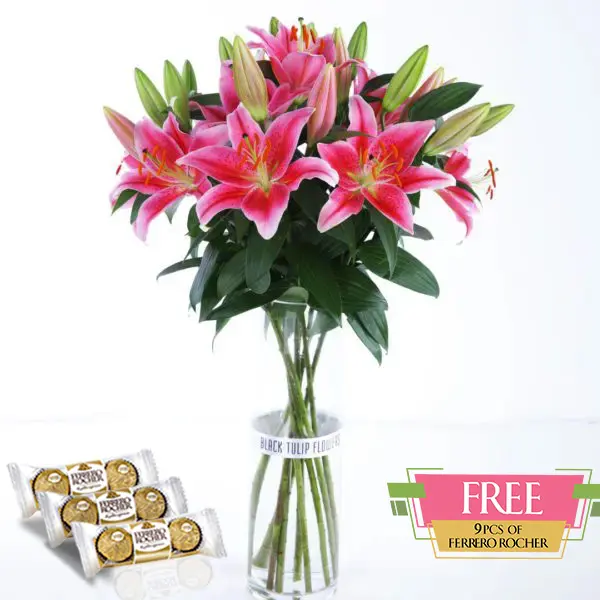 A bunch of pink lilies in a glass vase, accompanied by a box of Ferrero Rocher chocolates.