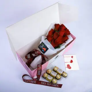 A pink gift box with a red ribbon, containing a bouquet of red roses and a box of Ferrero Rocher chocolates with a greeting card.