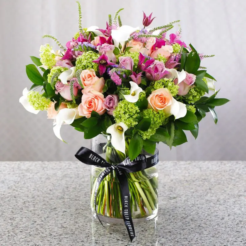 Lavish Love (1) Mix of fresh, vibrant flowers arranged in a clear glass vase tied with a black ribbon.