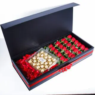 A black gift box with a red ribbon, with red roses and a clear box of Ferrero Rocher chocolates on a bed of red rose petals.