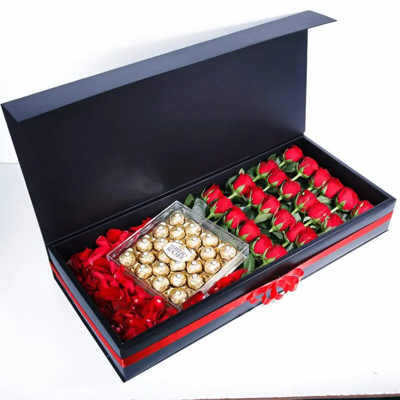 Romantic Trio with Ferrero (1) A black gift box with a red ribbon, with red roses and a clear box of Ferrero Rocher chocolates on a bed of red rose petals.