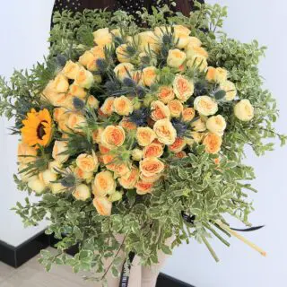 Vibrant yellow roses, blue thistles, and a sunflower with lush greenery all arranged in a lush, flowing design.