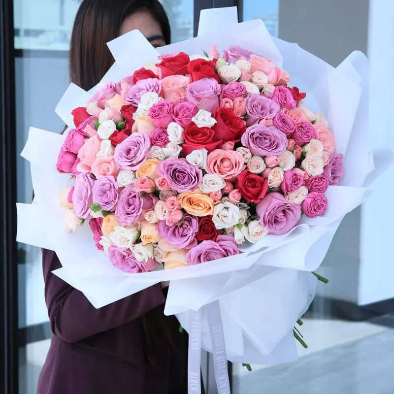 White and Mixed Emotion (1) A large mixed rose bouquet in red, pink, purple, orange, and white, wrapped in white paper and tied with a ribbon.