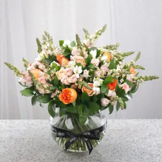 A clear glass vase featuring peach and orange roses, white snapdragons, and delicate blooms, tied with a black ribbon.