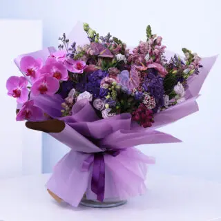 Bouquet of purple and pink flowers, including orchids, roses, and hydrangeas, wrapped in purple paper.
