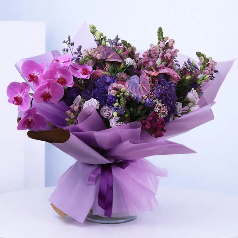 purple-royalty_2026 Bouquet of purple and pink flowers, including orchids, roses, and hydrangeas, wrapped in purple paper.