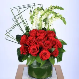 A floral arrangement in a glass vase, with red roses, white snapdragons, greenery, and geometric wire accents.