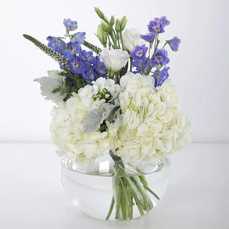 Pleasing Centerpiece (1) The centerpiece features blue delphinium, white hydrangeas, and other white flowers in a clear glass vase.