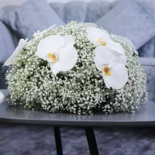 A large, round arrangement of white baby's breath with white orchids on top centerpiece, on a coffee table.