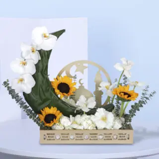 A floral arrangement with white orchids, sunflowers, and other flowers in a crescent moon shape, with a mosque cut-out in the background, in a light wooden box