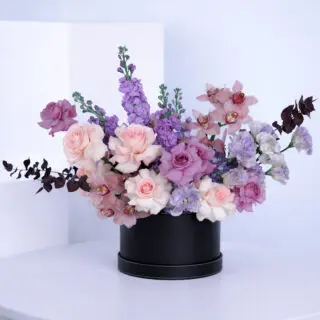 A luxurious mix of pink roses, purple roses, cymbidium, purple matthiola, purple lisianthus, blue hydrangeas, and eucalyptus, elegantly arranged in a sleek black box.
