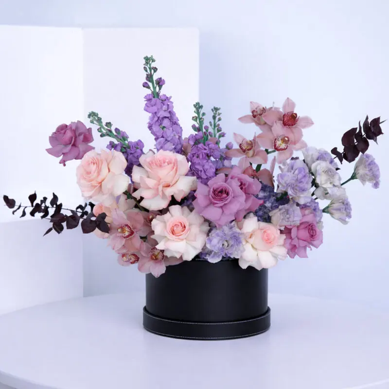 alluring-floral-box-2026 A luxurious mix of pink roses, purple roses, cymbidium, purple matthiola, purple lisianthus, blue hydrangeas, and eucalyptus, elegantly arranged in a sleek black box.