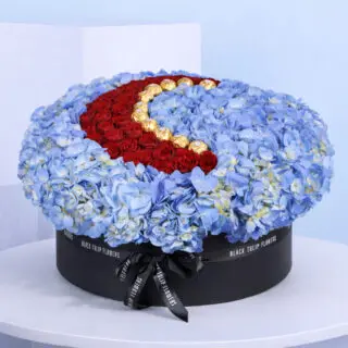 A round floral arrangement of blue hydrangeas, red roses, and gold chocolates, shaped like a crescent moon, in a black box with 'Black Tulip Flowers' branding