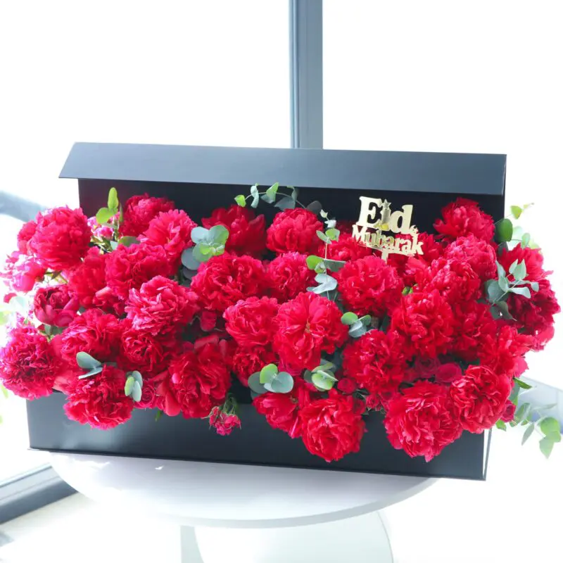 Eid Rejoice Flower Box (1) A black box filled with dark pink peonies and greenery. The lid of the box has "Eid Mubarak" written on it in gold.