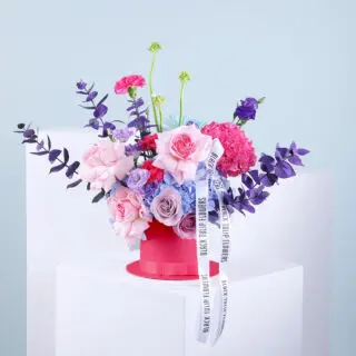 A mix of blue and pink hydrangeas, roses, lisianthus, carnations, ornithogalum, and chrysanthemum with eucalyptus in a pink hatbox tied with a white ribbon.