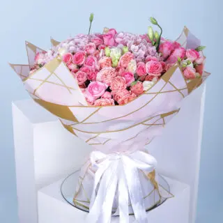 A bouquet of hydrangeas, baby roses, lisianthus, and pink roses wrapped in white with a gold border and a white ribbon.