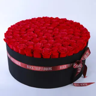 A sleek black circular box adorned with a red ribbon, filled with a stunning arrangement of vibrant red roses.