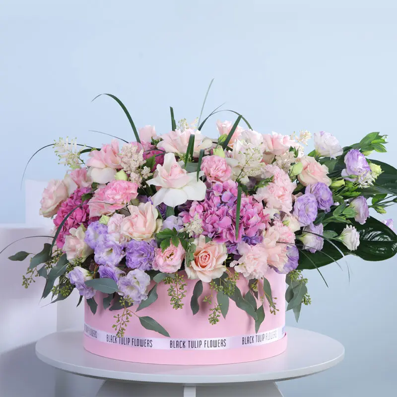 01 A large round arrangement of pink and purple roses, hydrangeas, lisianthus, and greenery in a pink box, tied with a white ribbon.