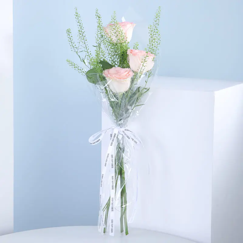A bouquet of pink roses wrapped in cellophane with a white ribbon, on a white table against a blue background.
