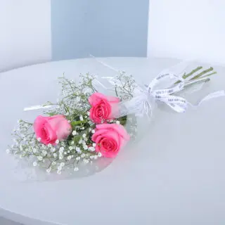 Small bouquet of 3 pink roses with baby's breath and greenery in a transparent wrap tied with white ribbon.