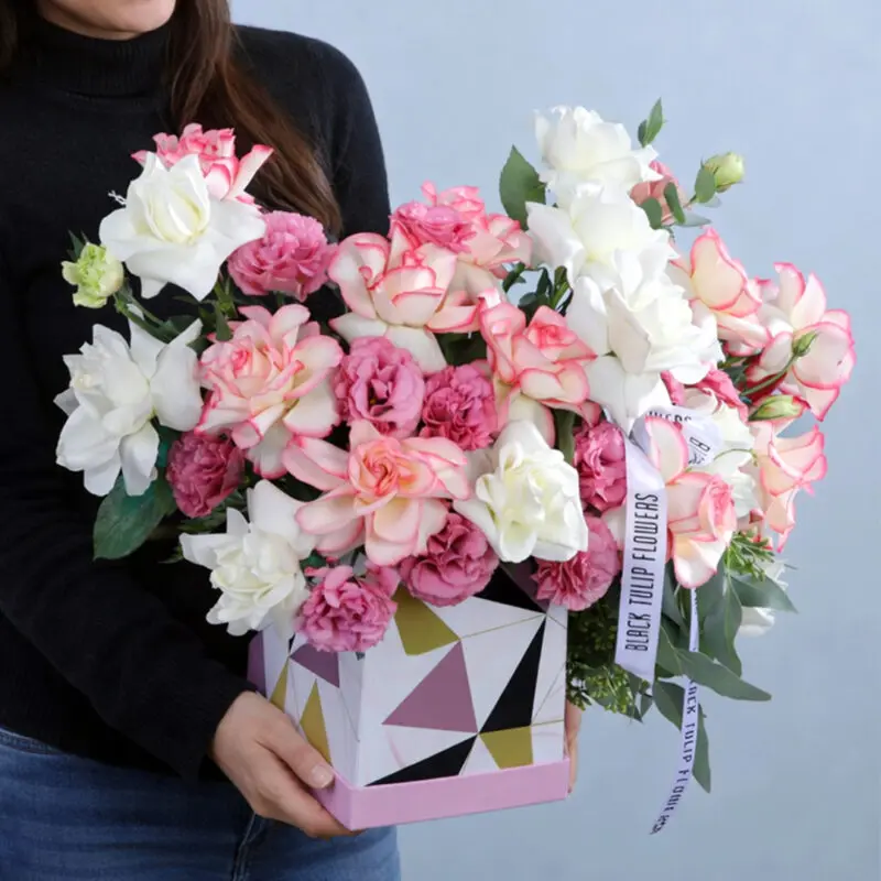 whispering-pastel-bloom-box_2026 A colourful floral arrangement in a geometric box with pink and white roses, with greenery accents adorned with a white ribbon.