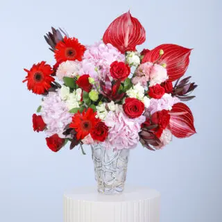 A floral arrangement in glass vase featuring red gerberas, pink hydrangeas, red roses, and striking anthurium leaves.
