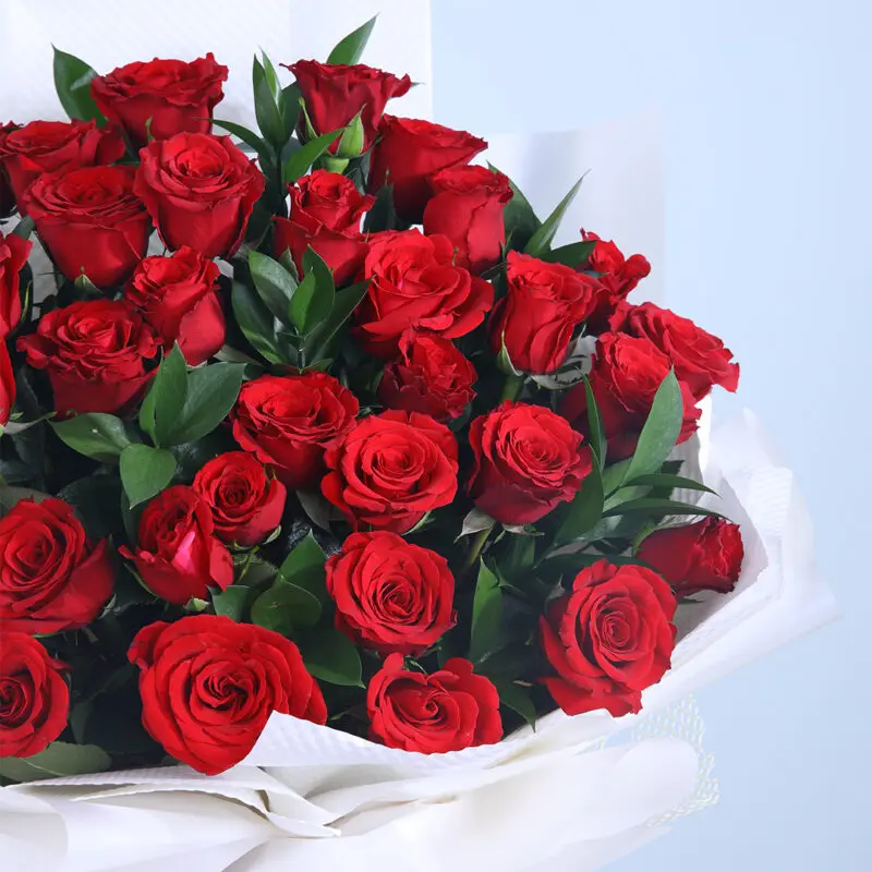 Bouquet featuring 30 red roses wrapped in white, adorned with fresh green leaves and tied with a maroon ribbon.