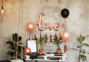A decorated room with a "love" balloon, other balloons, streamers, plants, and a blank photo frame. The room has a rustic, industrial feel with exposed brick walls and a hanging swing