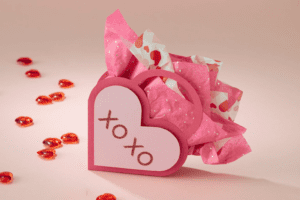 A pink heart-shaped gift box with a cutout design that spells out "XOXO." The box is filled with pink tissue paper and surrounded by small red heart-shaped candies.