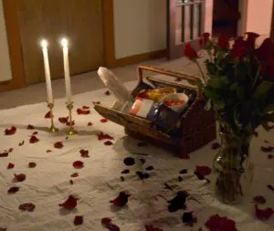 A romantic indoor picnic scene with a wicker basket filled with food, a vase of red roses, two lit candles, and scattered rose petals on a white sheet.