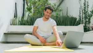 A man sitting on a yoga mat, using a laptop to participate in a virtual yoga class with a boyfriend.