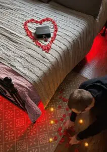 A person decorating the room with rose petals for the scavenger hunt. A black box with a red lock is placed on the bed inside a heart shape formed with rose petals