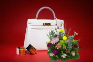 A white Hermes Kelly 28 handbag, paired with two orange Hermes bracelets and a vibrant bouquet of flowers. The background is a solid red color.