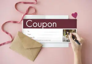 A blank coupon with the word "Coupon" written in bold on top. There are lines for "To," "From," and "Voucher ID," along with a small image of a dinner table setting. A hand is holding a pen above the coupon, ready to write on it. A pink ribbon and a paper heart are also visible in the image.