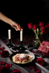 A romantic Valentine's Day dinner scene with a plate of spaghetti covered in red sauce and grated cheese, lit candles, a bouquet of red roses, and rose petals scattered on the table
