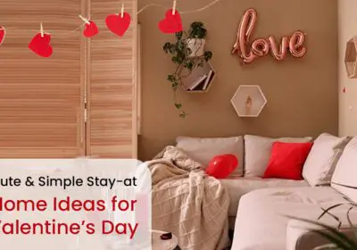 A cozy living room decorated for Valentine's Day. There is a sofa with red pillows, a string of heart-shaped decorations, a "Love" balloon, and other Valentine's Day decor. The text "Cute & Simple Stay-at-Home Ideas for Valentine's Day" is also included.