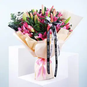 A bouquet of pink lilies wrapped in beige paper with a pink ribbon and black ribbon accents.