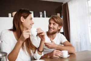 A young, smiling couple sits at a kitchen table, enjoying their morning coffee