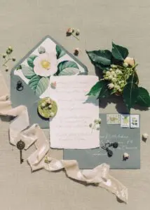 A flat lay image showcasing a vintage-inspired wedding invitation suite. The invitations are on textured paper with handwritten calligraphy. There are also envelopes, stamps, a velvet ribbon, a key, and floral accents, including a camellia flower and berries. The overall aesthetic is elegant and romantic.