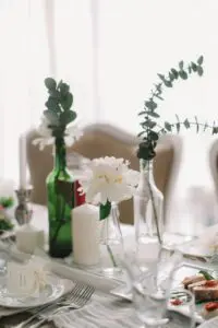 A beautifully set table with a minimalist, elegant aesthetic. White peonies and eucalyptus sprigs are arranged in recycled glass bottles as centerpieces. The table is set with white plates, silverware, and wine glasses. There are place cards with names written on them, adding a personal touch.