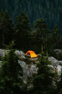A bright orange tent pitched on a rocky ledge surrounded by a dense forest. The dark, towering trees create a dramatic contrast with the vibrant tent.