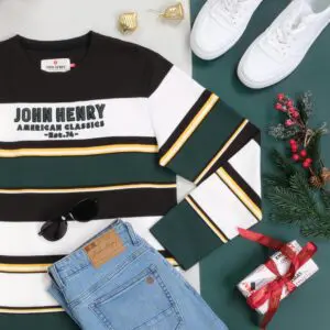 A flat lay showcasing a John Henry striped sweater, jeans, sunglasses, sneakers, and a gift box with a red ribbon. The image has a festive feel with Christmas decorations like a pine branch and ornaments.