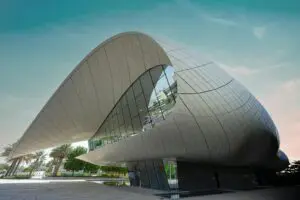 A modern, white building with a unique, flowing design, reminiscent of a seashell. The building features large glass windows and a curved roof. Palm trees and a blue sky with clouds complete the scene.