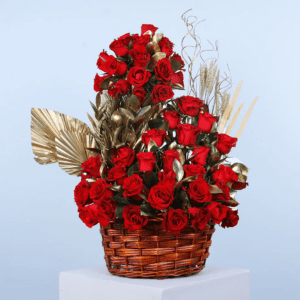 A large, luxurious flower arrangement in a wicker basket. The arrangement features a towering display of red roses, accented with gold leaves and wheat stalks.