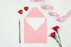 A pink envelope with a blank card inside, next to a pen, heart-shaped chocolates, and carnations.