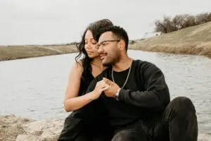 A couple sitting on a rock by a lake, enjoying a scenic view. The man is wearing a blue shirt and the woman is wearing a red shirt. Both are looking at the view in front of them.